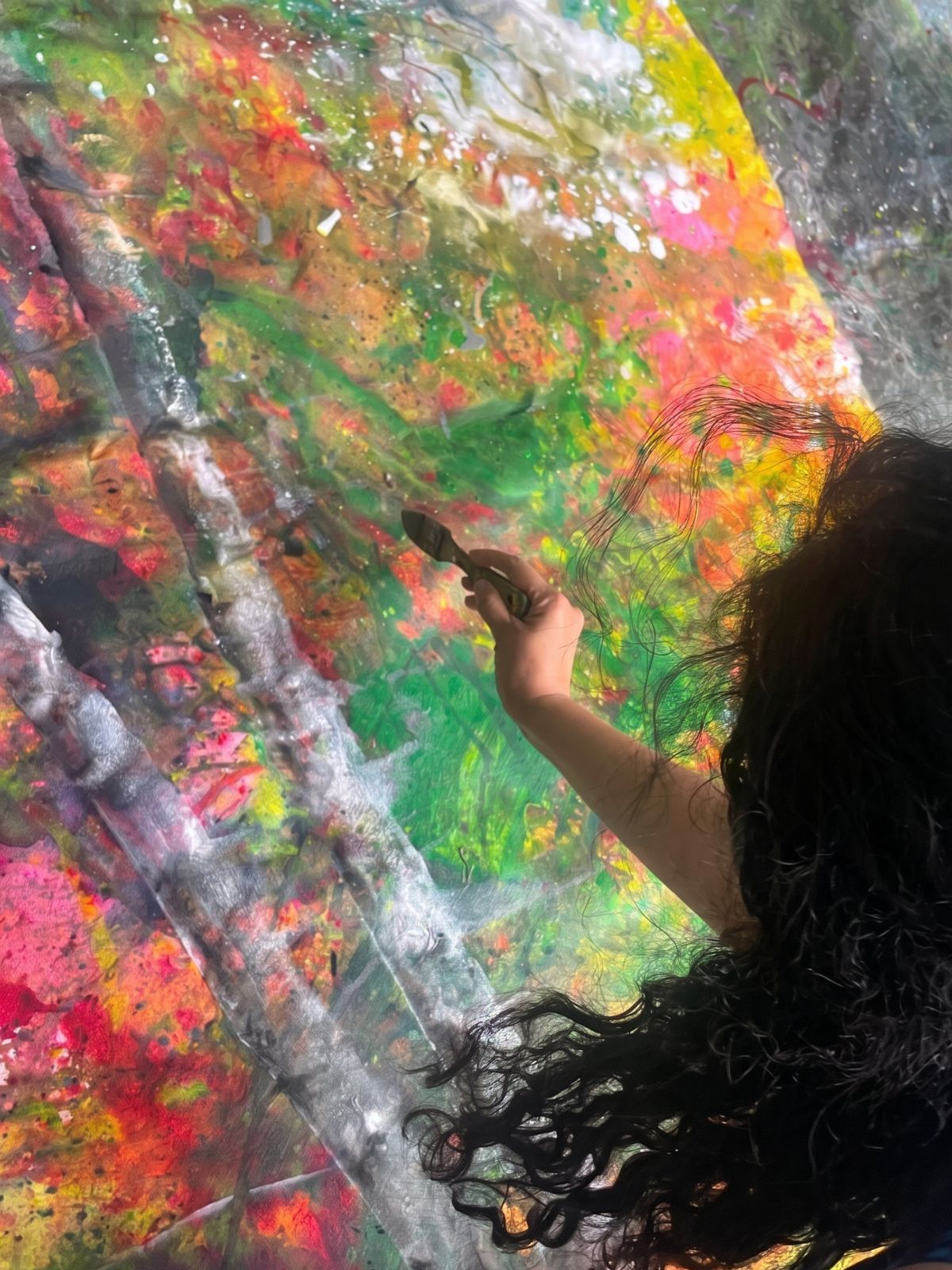 A workshop participant painting on a vertical plastic sheet — vivid pink, green, yellow and orange splatters worked across the surface, hand visible mid-stroke.