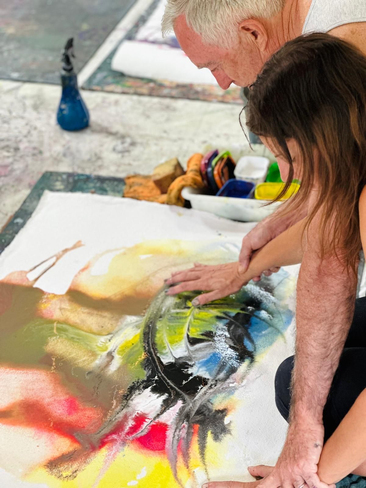 Two workshop participants painting together on a shared canvas, hands working side by side.