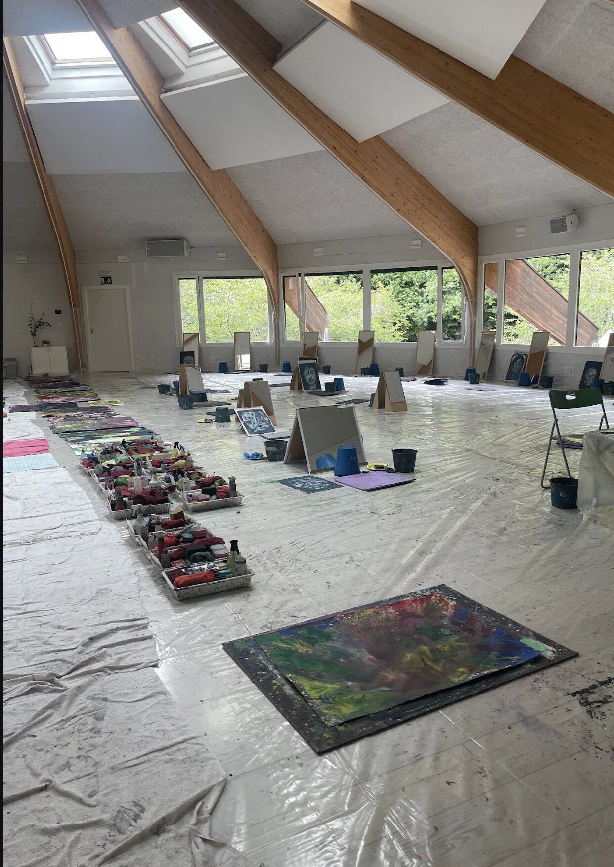 The empty Amalurra workshop hall before a session — wooden beams, skylights, plastic floor sheeting, easels and canvases laid out.