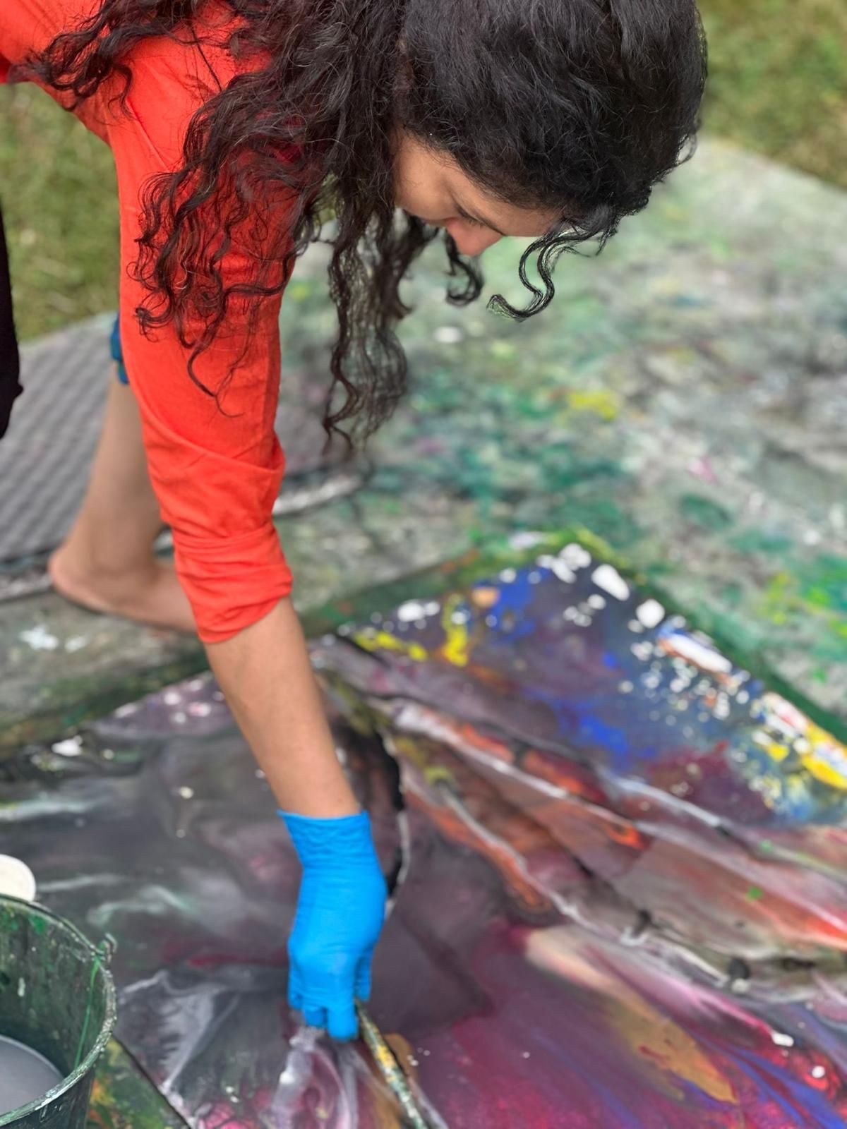 A workshop participant in a red top and blue glove painting on a glossy dark canvas with magenta, orange, and blue passages.