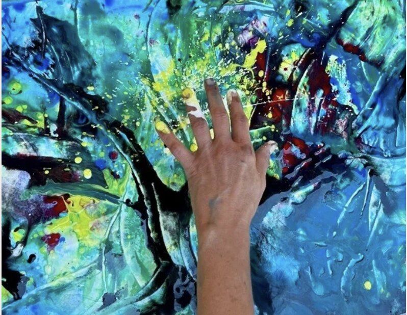 A close-up of a hand pressed onto a vivid abstract painting — turquoise, yellow, and red splatters with paint smears on the fingers.