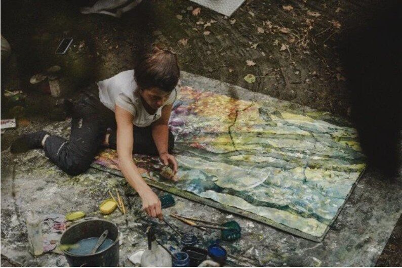 A workshop participant kneeling on a tarpaulin painting a large abstract canvas in greens and yellows, brushes and paint trays beside her.
