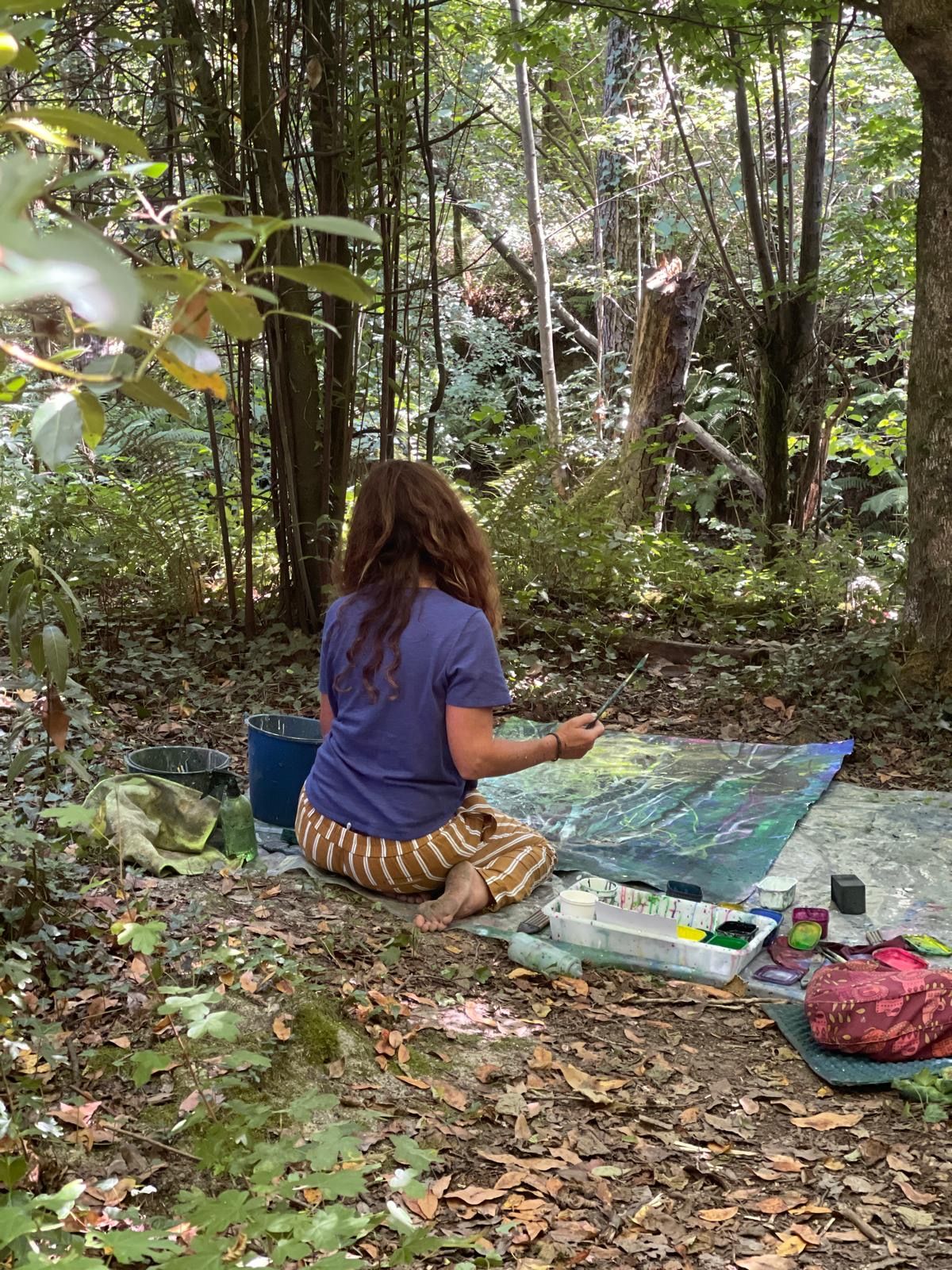 A workshop participant in a blue T-shirt and striped trousers seated cross-legged painting in a fern-filled forest clearing.