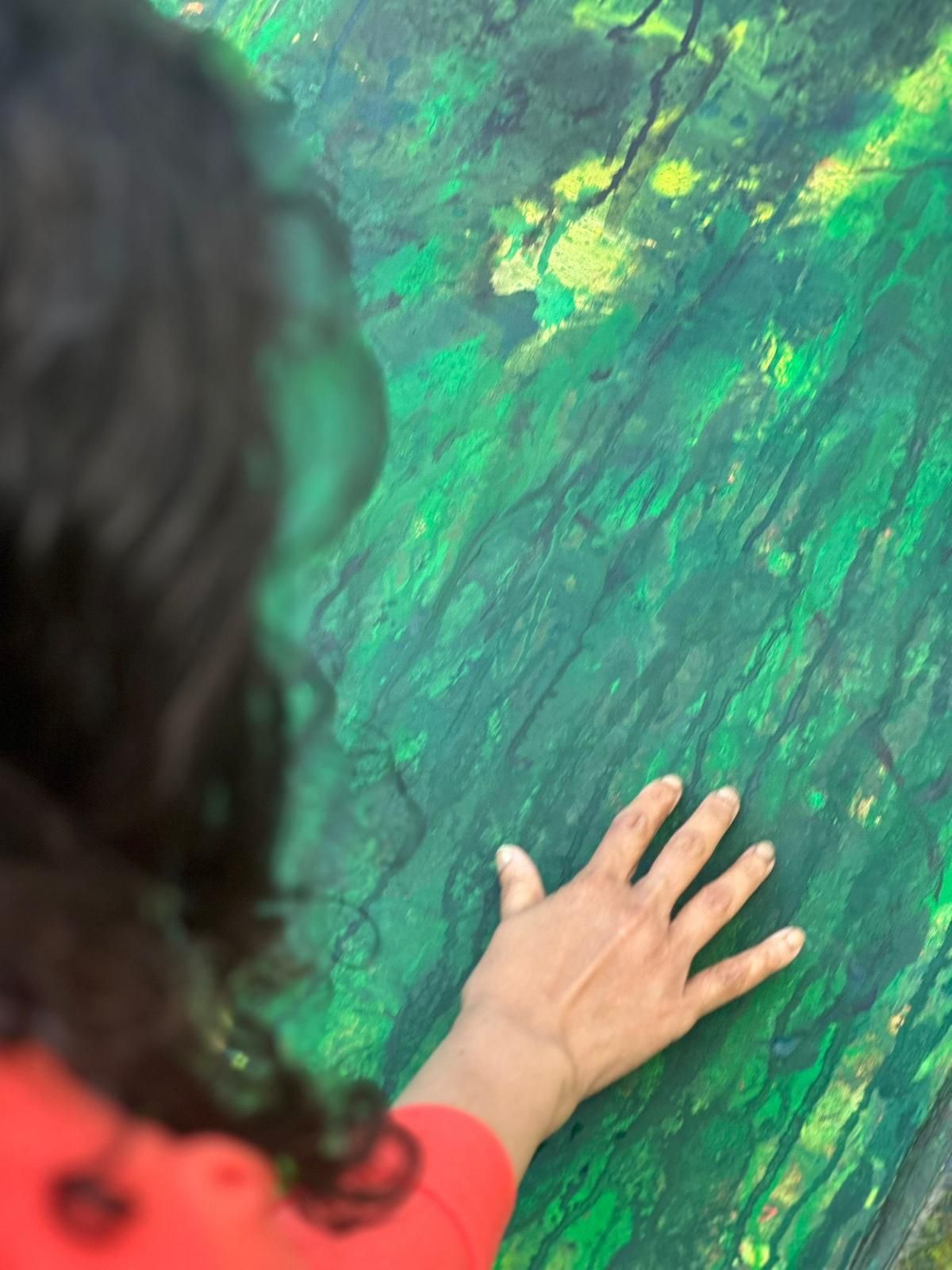 A close-up of a hand pressed onto a deep-green textured painting with flashes of yellow — long dark hair edges the frame.
