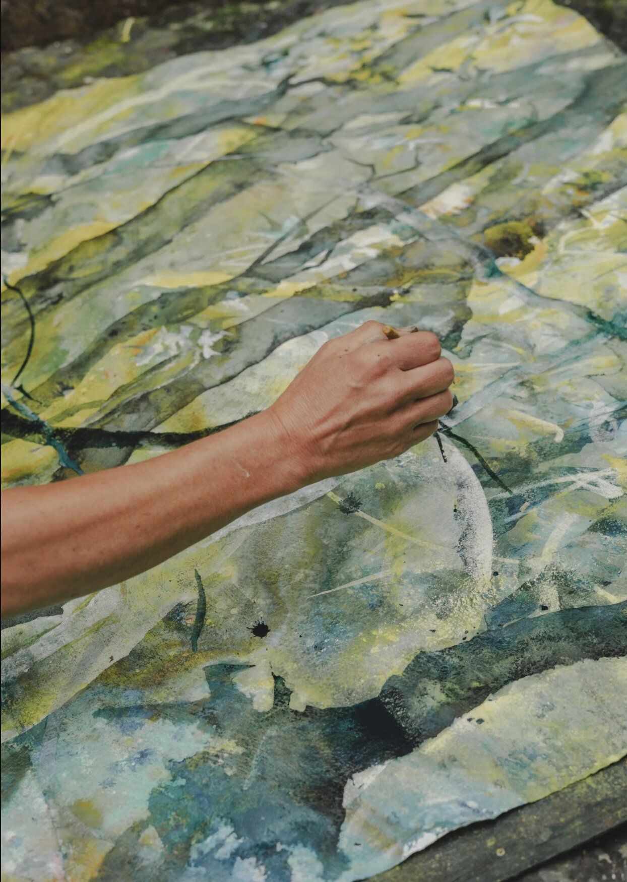 A close-up of an arm reaching across a wrinkled, pale watercolour painting in yellows and soft greens — water-marks and veining across the paper.