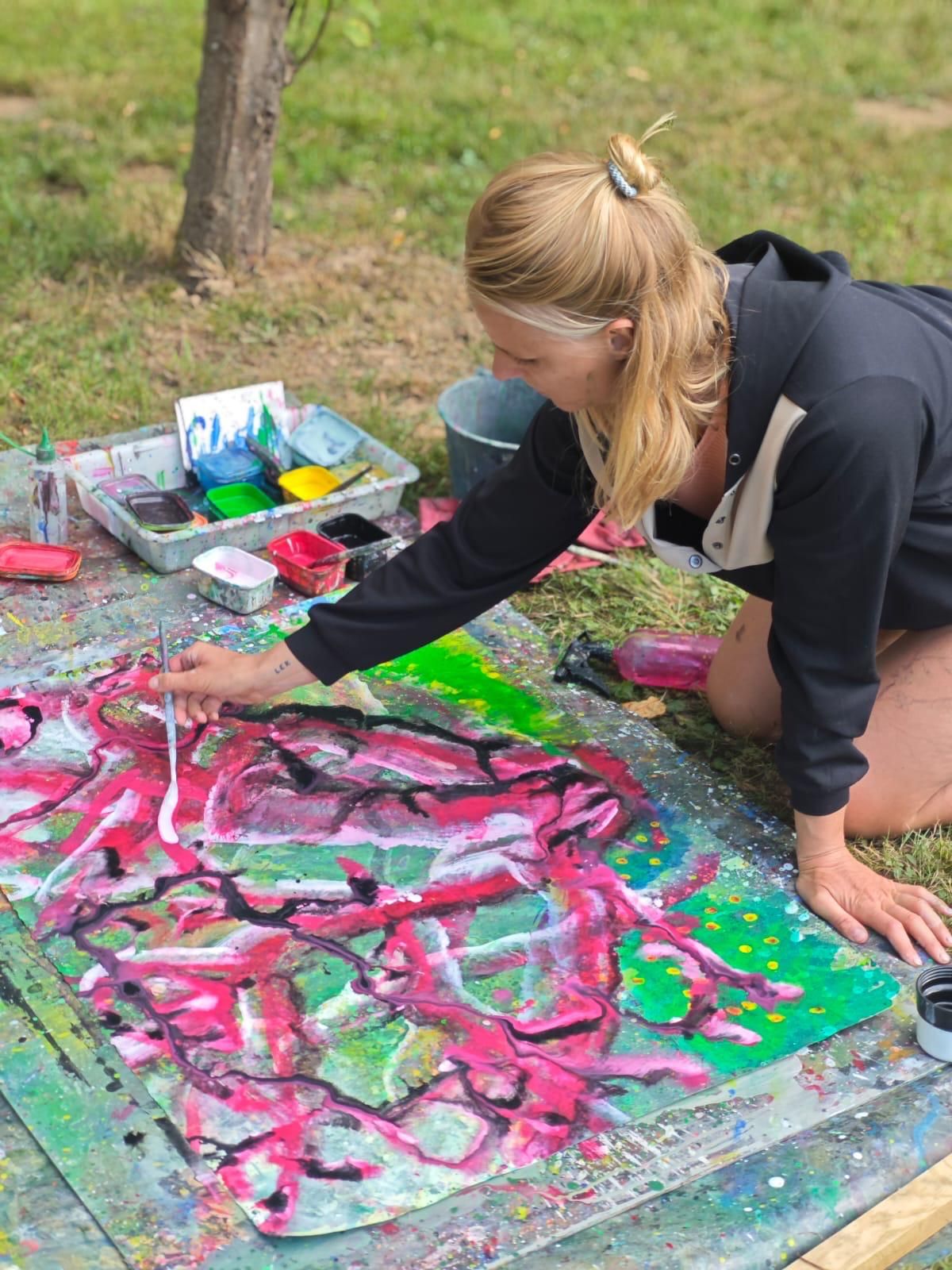 A workshop participant outdoors painting in vivid pink and green, blonde hair tied back.