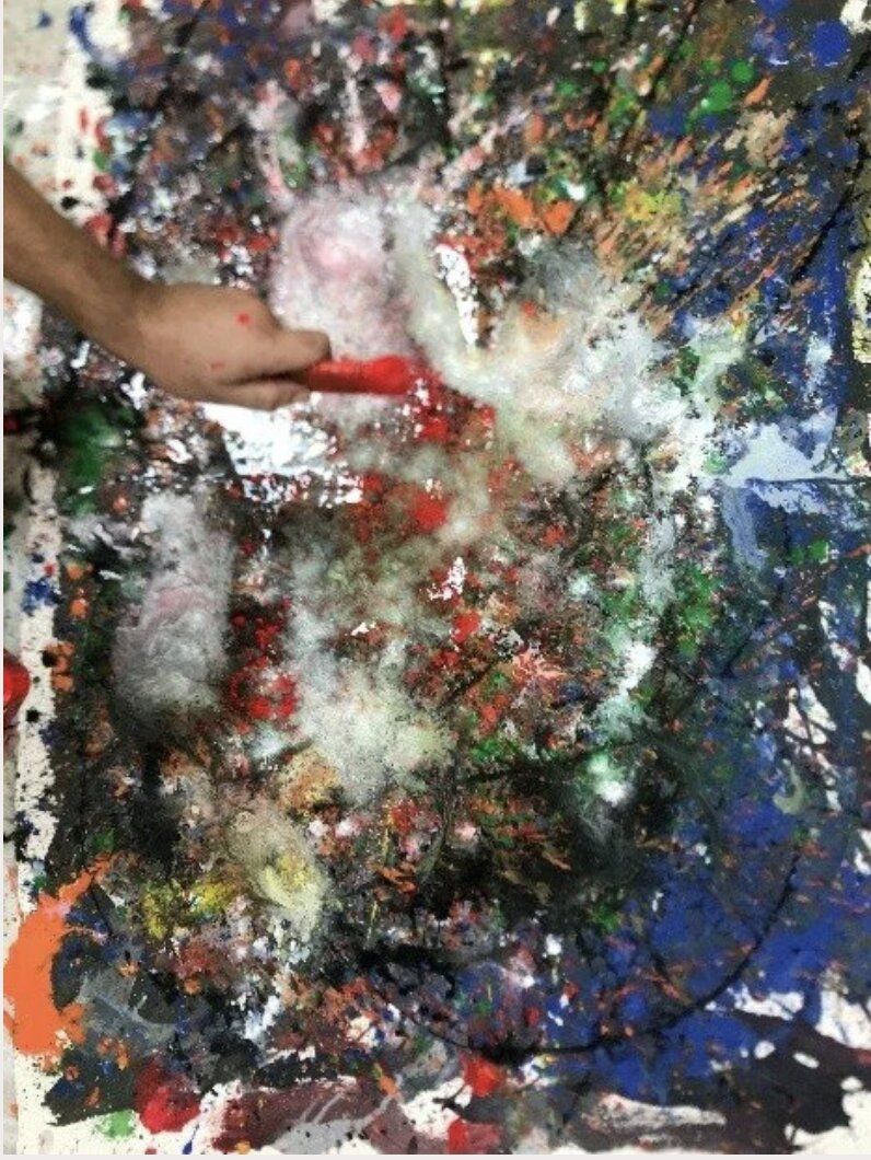 A close-up of a hand with a red-tipped brush working on a chaotic painting — blues, greens, reds, and white feathery textures.