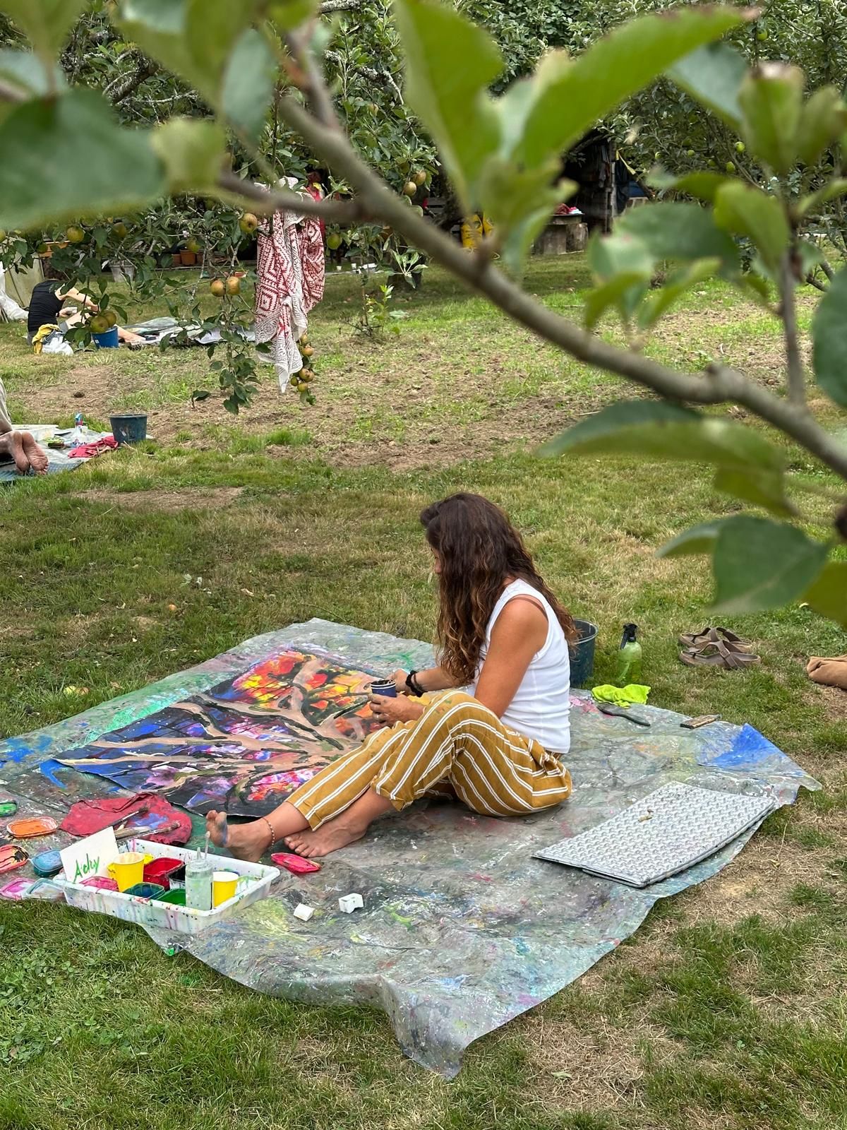 A workshop participant seated and painting under fruit trees, relaxed posture.