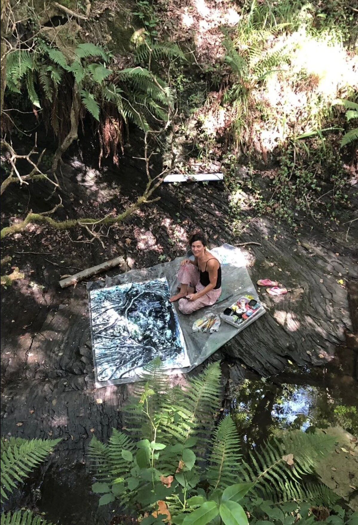 A workshop participant seated on rocks beside a forest stream, painting a dark abstract canvas, ferns and dappled light all around.