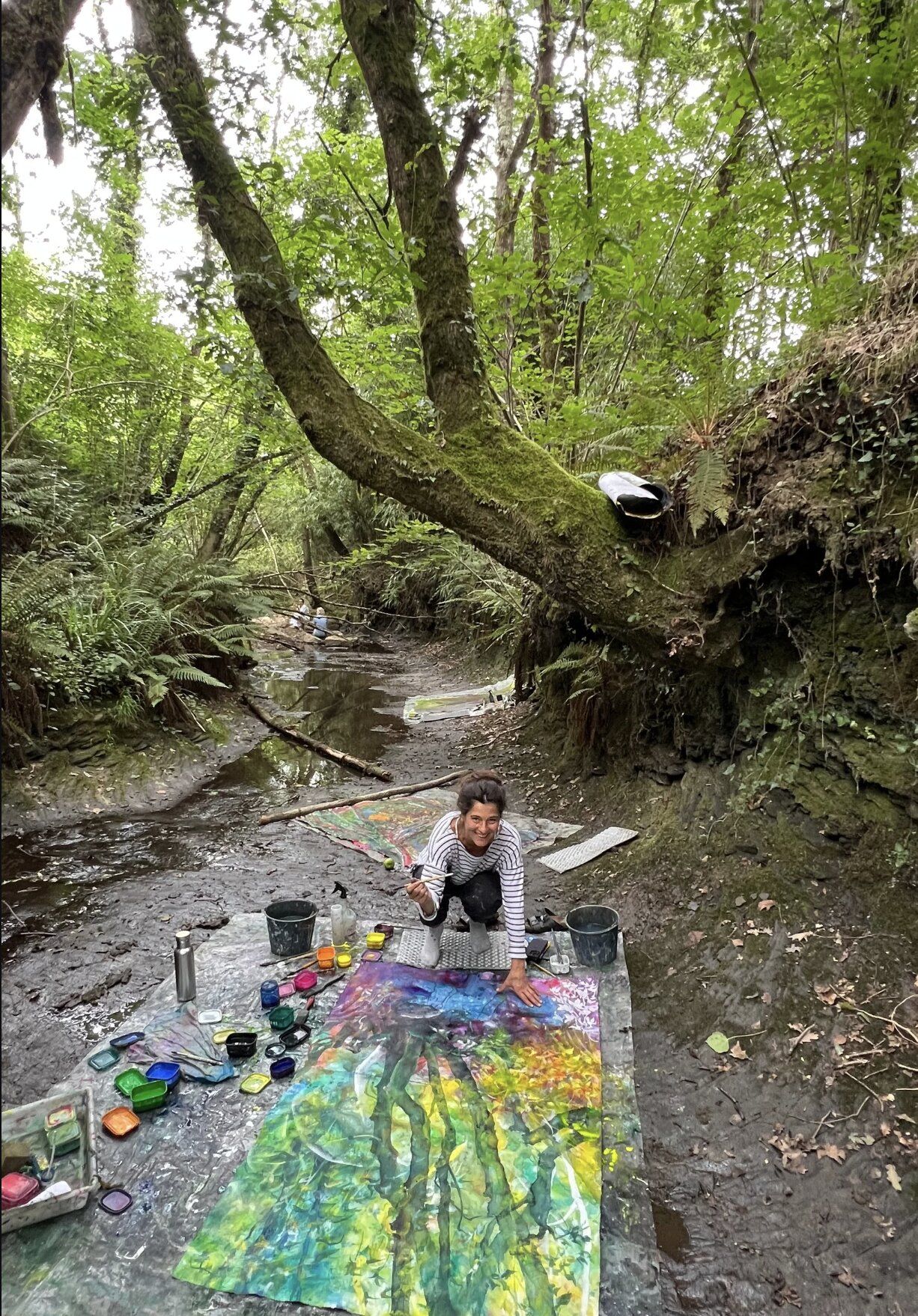 A workshop participant in a striped shirt crouching in a muddy forest stream, smiling, painting a colourful canvas under overhanging branches.
