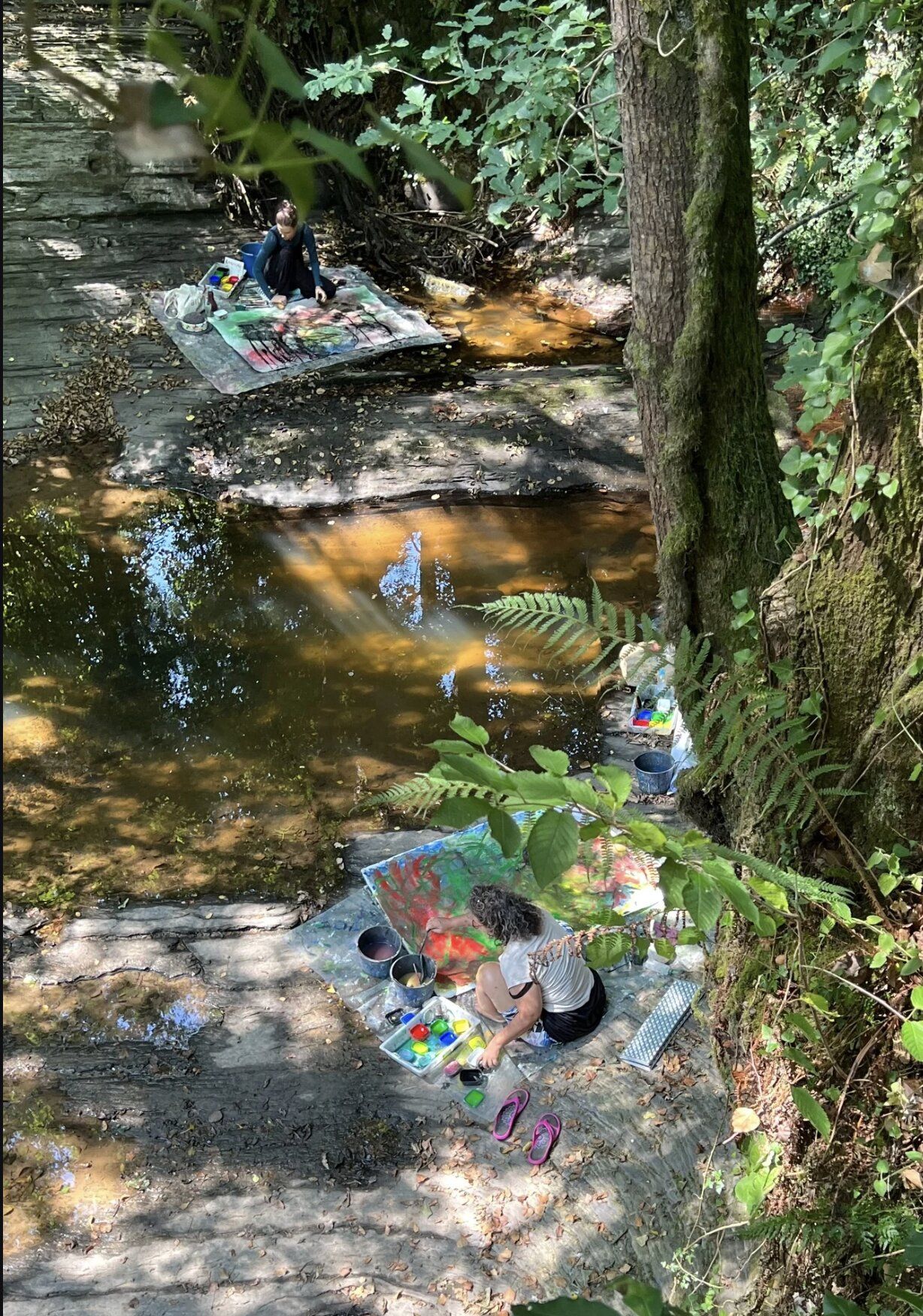Two workshop participants painting on tarpaulins in a shallow forest stream bed — ferns, mossy banks, dappled sunlight.