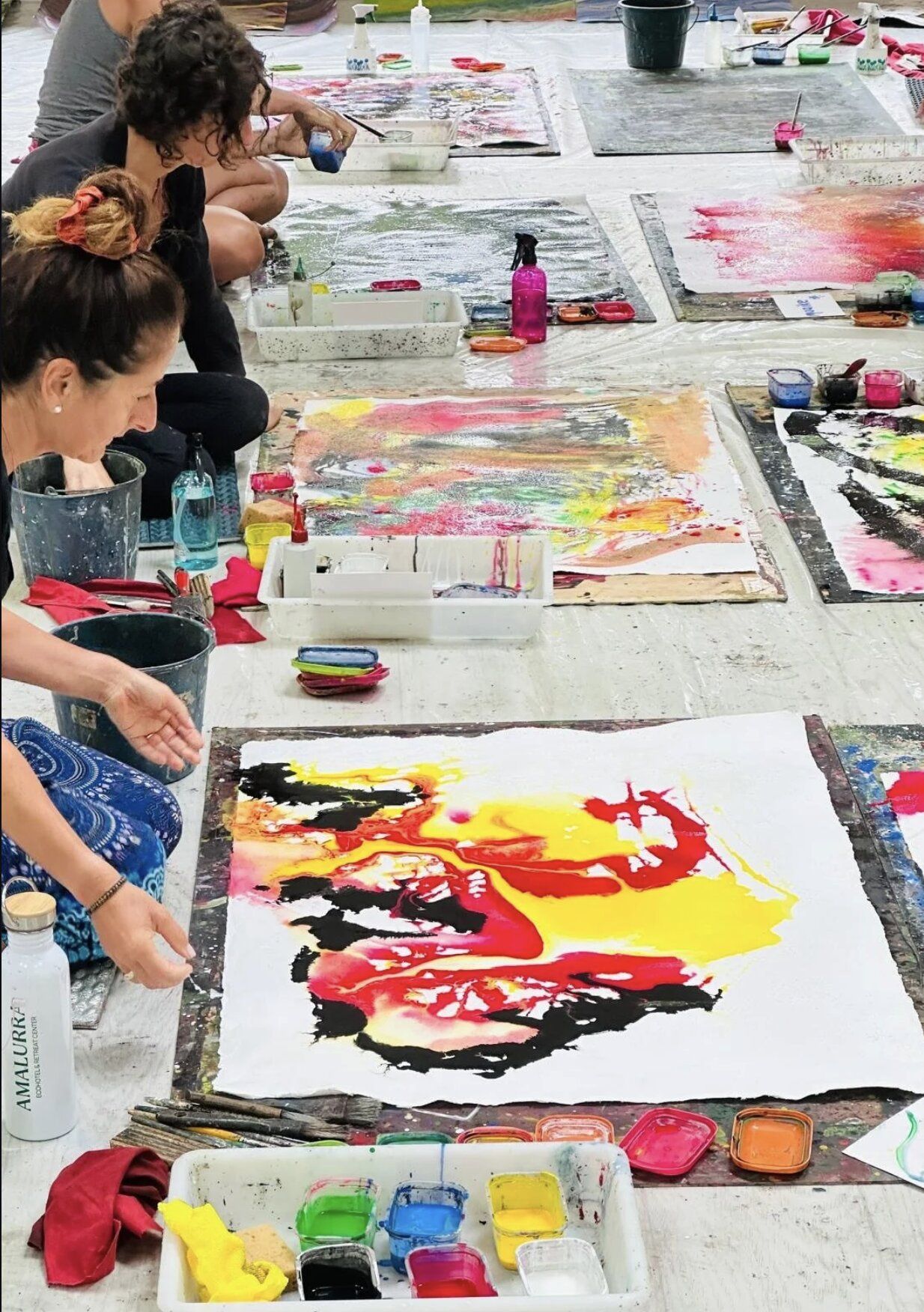 Workshop participants kneeling beside their canvases — a dramatic painting in yellow, red and black in the foreground, paints and brushes scattered.